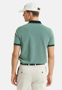 Camisa polo verde de manga corta con collar azul marino y detalles a rayas, combinada con pantalones de color claro y una gorra blanca, vista desde la parte de atrás.