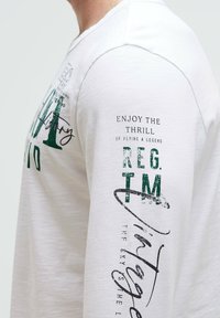Long-sleeve, cream cotton T-shirt featuring green text and black accents with varied font styles and prints on the front and right sleeve.
