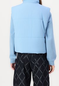 Helle blaue Steppweste mit hohem Kragen, gestepptem Abschnitt und gerippten Bündchen; kombiniert mit dunklen gemusterten Hosen mit einem Rautenmuster.