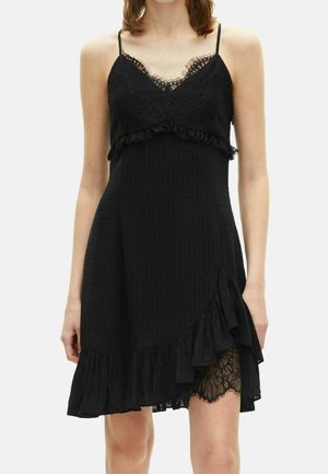 Kobieta w czarnej sukience bez rękawów z koronkowymi zdobieniami na dekolcie i krawędzi sukienki, z falbankami i asymetrycznym dołem.