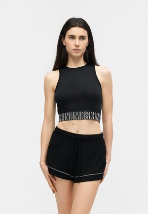 Femme aux longs cheveux bruns portant un crop top noir sans manches avec une bande logo Moschino blanche et un short noir à bordure blanche.