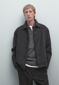 Gray wool blend jacket with a collar, front buttons, and two side pockets, layered over a gray sweater and white shirt, with dark pants.