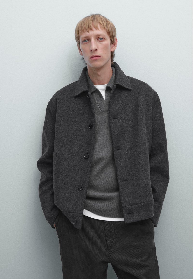 Gray wool blend jacket with a collar, front buttons, and two side pockets, layered over a gray sweater and white shirt, with dark pants.