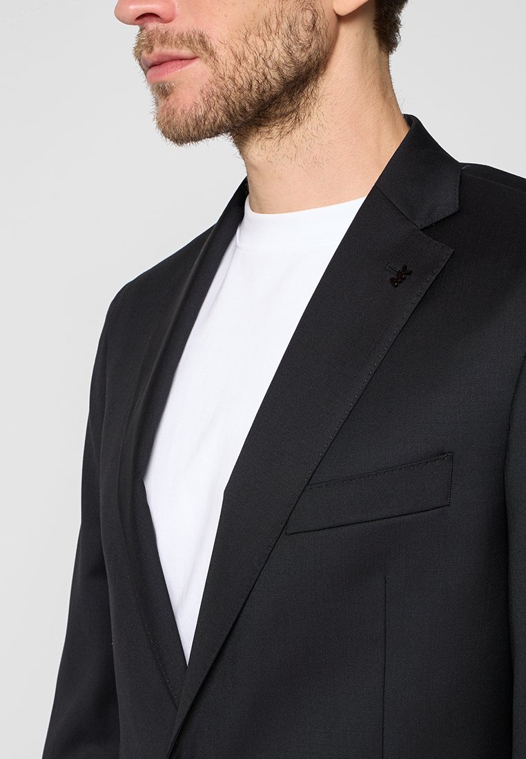 Een bebaarde man in een zwarte blazer over een wit T-shirt, weergegeven van borst tot kin tegen een eentonige achtergrond.