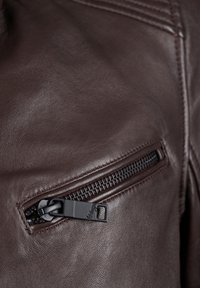 Bruine leren jas met een borstzak met ritssluiting en zwarte rits. Textuur met zichtbare stikdetails.