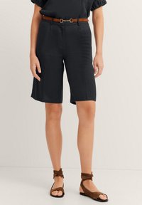 Schwarze Bermuda-Shorts mit einem entspannten Schnitt, ausgestattet mit einem Bund. Kombiniert mit braunen Sandalen mit Knöchelriemen und einem passenden Gürtel. Flache Textur.