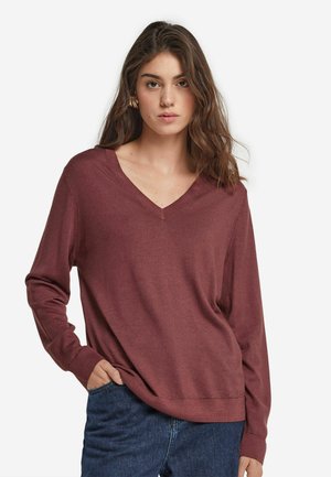 V-ausschnitt weinrot Pullover mit langen Ärmeln, weichem Material und lockerer Passform, kombiniert mit blauen Jeans, Hände in den Taschen.