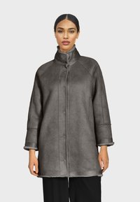 Sivi kaput od umjetnog janjećeg krzna s visokim ovratnikom, dugim rukavima i vidljivim šavovima. Meki materijal i ravna silueta.