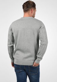 Pull en tricot gris avec des poignets côtelés et un design en cable texturé sur les manches. Porté avec un jean bleu, mettant en valeur une silhouette ajustée.