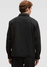 Mustanud, pika varrukaga jakk kraega, sileda tekstuuriga ja minimaalsete õmblusdetailidega. Tagantvaates on näha kõrge mugavusaste ja küljetaskud.