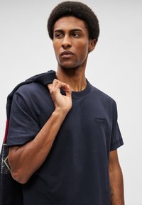 Μπλέ βαθύ βαμβακερό μπλουζάκι με στρογγυλή λαιμόκοψη και κοντά μανίκια, που διαθέτει μια μικρή ετικέτα λογότυπουστο στήθος και ένα υφασμάτινο πουλόβερ ριγμένο στον ώμο.