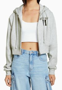Felpa corta grigia con zip, tasca frontale e cappuccio regolabile, con la scritta "Melbourne" e una lettera "M" in nero.