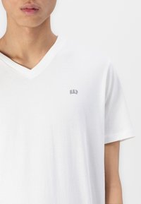 T-shirt blanc à col en V en coton doux, avec un petit logo "GAP" gris sur la partie supérieure gauche de la poitrine. Texture lisse, manches courtes.