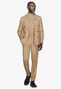 Beige double-breasted jakkesæt med en tætsiddende pasform, parret med en matchende turtleneck og mørkebrune sko med teksturerede detaljer.