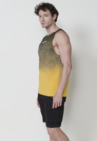 Siva in rumeni gradient majica brez rokavov s krožnim izrezom. Črne kratke hlače s žepi. Model stoji v profilu.