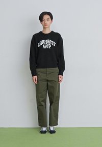 Persona stāv pretī baltai sieni, ģērbusies melnā Carhartt WIP džemperī, olīvu zaļos biksēs un melnbaltās sporta apavā uz zaļa grīdas.