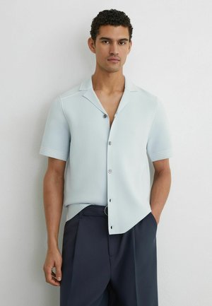 Mand med kort krøllet hår iført lyseblå kortærmet skjorte og mørkeblå bukser, stående op ad en hvid væg.