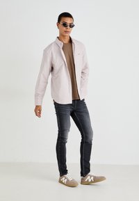 Striped button-up shirt in white and light pink, brown t-shirt, dark jeans, and beige sneakers with white accents. Sleek sunglasses.