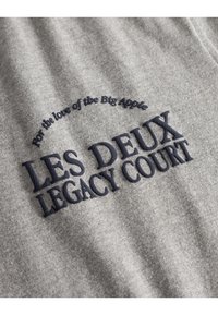 Pilkas audinys su tamsiai mėlynai išsiuvinėtu tekstu: "LES DEUX LEGACY COURT" ir aukščiau jo: "Dėl didžiosios Obuolio meilės".