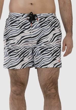 Mann trägt schwarz-weiße, zebra-gestreifte Badeshorts mit schwarzer Kordel auf hellgrauem Hintergrund.