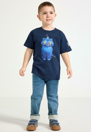 Ung dreng står iført en mørkeblå t-shirt med en blå tegneserie kat, lyseblå jeans med opbrettede bukseben og brune sneakers med blå snørebånd.