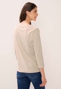 Frau mit langen dunklen Haaren, die ein beiges geripptes Langarmoberteil und blaue Jeans trägt, schaut nach rechts vor einem schlichten hellen Hintergrund.