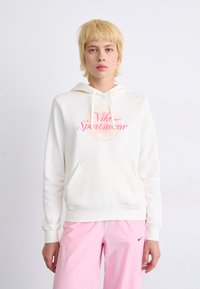 Balta hoodie su priekiu kišene, raišteliu ir rožiniu spaudiniu "Nike Sportswear" ant krūtinės. Derinama su šviesiai rožinėmis kelnėmis.