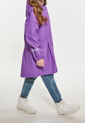 Enfant marchant portant un imperméable violet avec une bande réfléchissante, un jean bleu et des baskets blanches montantes sur un fond neutre.