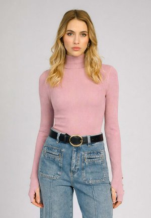Femme aux cheveux blonds portant un pull à col roulé côtelé rose et un jean taille haute bleu avec de grandes poches avant et une ceinture noire.