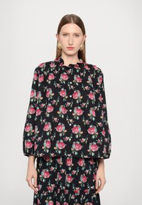 Svart bluse med livlig rosa og teal blomstermønster, volangkrage, knappelukking foran og lange ermer. Glatt stoff med avslappet passform.