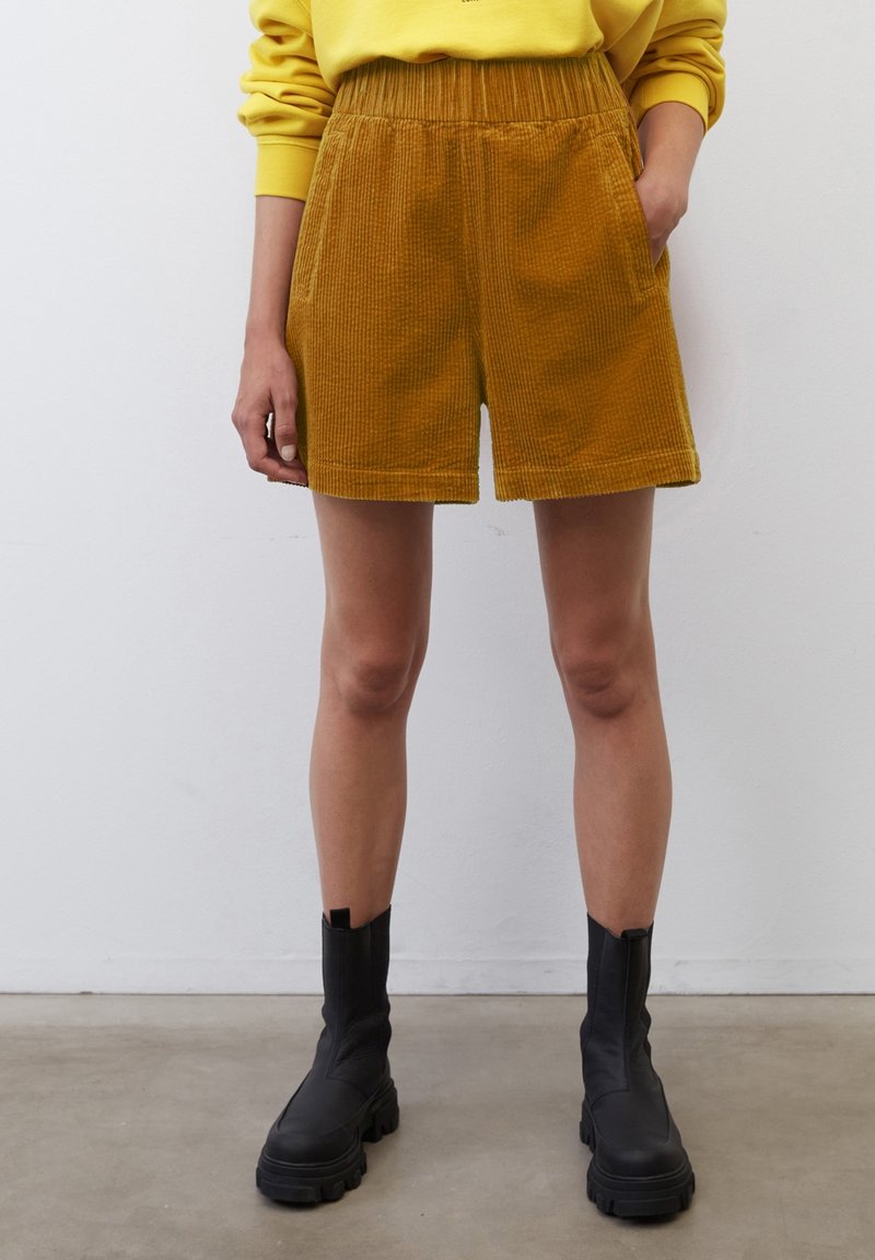 Personne portant un short en velours côtelé jaune moutarde, un sweat-shirt jaune et des bottines noires épaisses, debout devant un mur blanc uni.