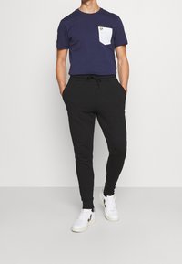 Marineblaue Baumwoll-T-Shirt mit einer weißen Tasche und kleinem Logo, kombiniert mit schwarzen Jogginghosen. Weiße Sneakers mit schwarzen Akzenten. Cropped-Schnitt, elastischer Bund.