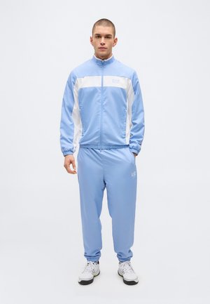 Moški v svetlo modri in beli trenirki EA7 s ujemajočimi hlačami ter belimi supergami, ki stoji pred enobarvnim ozadjem.