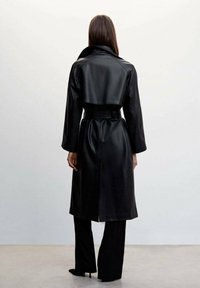 Manteau trench-coat en cuir noir avec une ceinture à la taille, des manches longues et une fente à l'arrière pour faciliter les mouvements. Il présente un large col et une texture lisse.