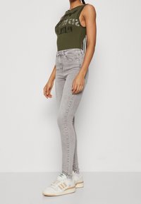 Jeans ajustados de talle alto grises combinados con una camiseta de tirantes verde con textura y estampado gráfico; llevados con zapatillas deportivas blancas y beiges.