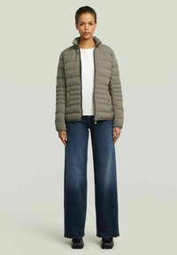Vrouw staat in een licht olijfgroene gewatteerde jas over een witte top, donkere wijde jeans en zwarte rechthoekige schoenen.