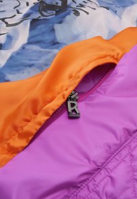 Colorful jacket with orange and purple panels, featuring a zippered pocket with a black metal pull. Smooth, shiny fabric texture.