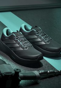 Paire de chaussures de course noires avec trois bandes, empeigne Primeknit et logo Mercedes-Benz sur la languette, posées sur un sol sombre près du matériel d’exercice.