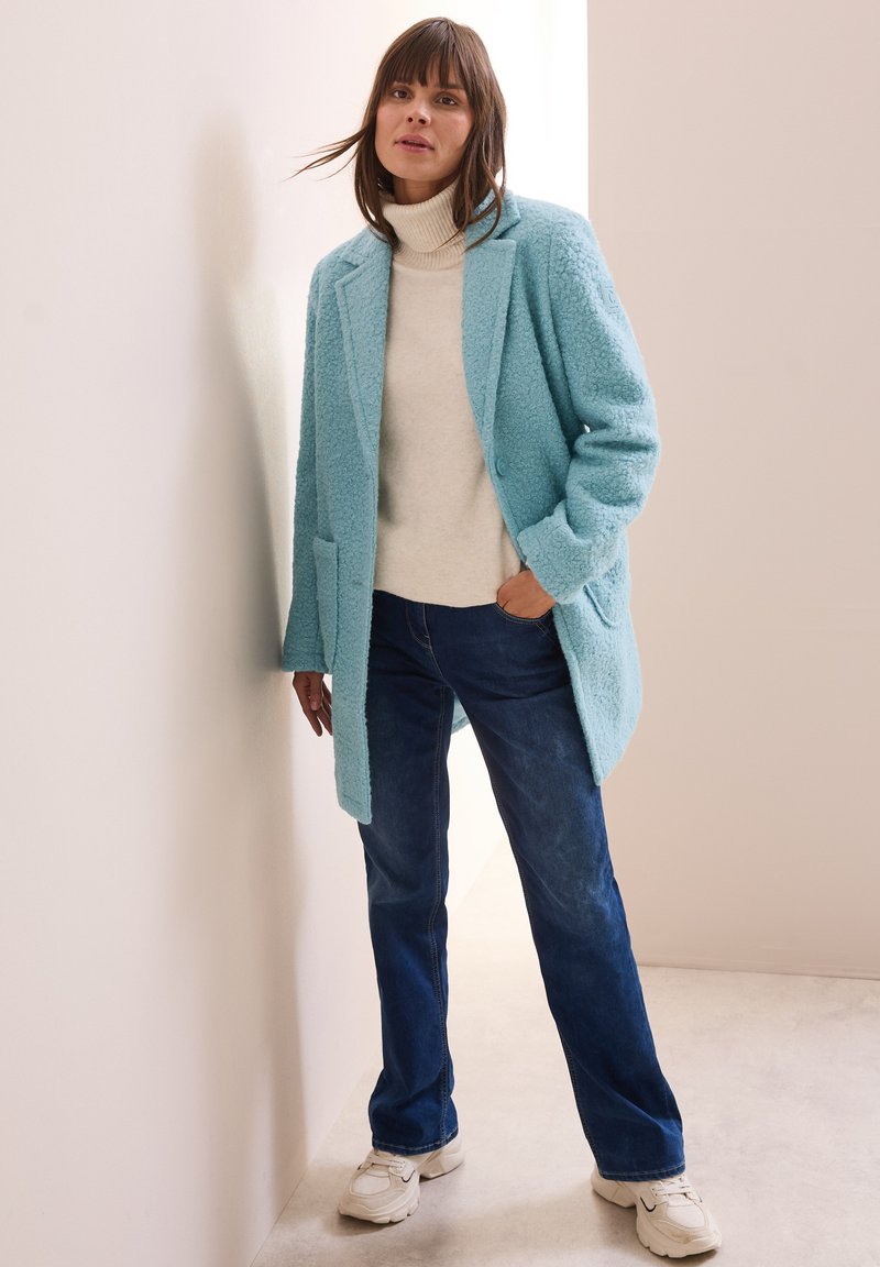 Cappotto turchese con texture, dal design sartoriale, dotato di due tasche frontali, indossato sopra un maglione a collo alto color crema e jeans blu, abbinato a sneakers bianche.