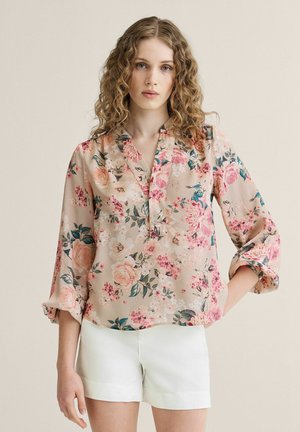 Ung kvinde med krøllet hår iført en beige blomsterbluse med opsmøgede ærmer og hvide shorts, stående mod en ensfarvet baggrund.