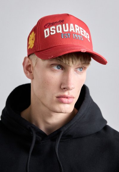 Röd basebollkeps med vit och blå "DSQUARED2"-text, etableringsåret 1995 och en gul lejonemblem.