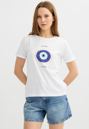 Joven mujer que lleva una camiseta blanca con un gráfico de ojo azul y el texto "power inside", combinada con shorts vaqueros de color azul claro, de pie con las manos detrás de la espalda.