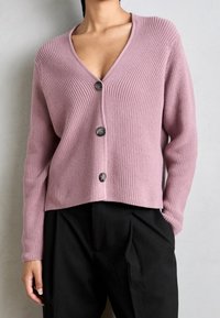 Jasnoróżowy kardigan z prążkowanego materiału, z dekoltem w kształcie litery V i trzema guzikami na przodzie. Ma długie rękawy i krótki krój. Widoczne czarne spodnie.