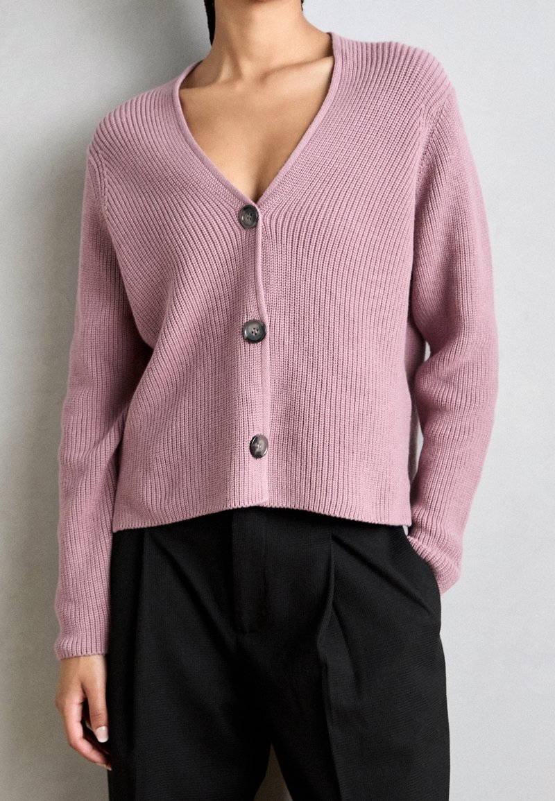 Jasnoróżowy kardigan z prążkowanego materiału, z dekoltem w kształcie litery V i trzema guzikami na przodzie. Ma długie rękawy i krótki krój. Widoczne czarne spodnie.
