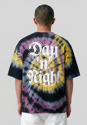 Persoon met kort haar die een zwart, geel en paars tie-dye T-shirt draagt met de tekst "Day n Night" op de achterkant, staand tegen een effen achtergrond.