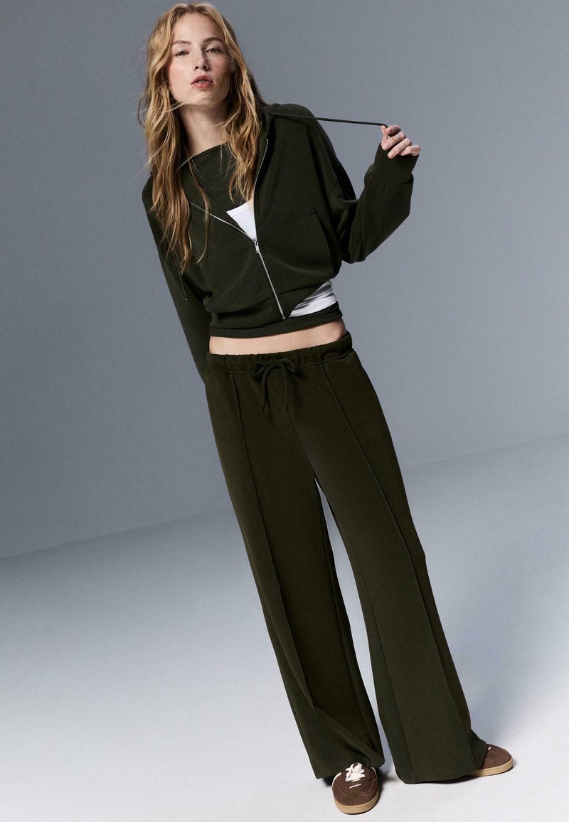 Dunkelgrüner Hoodie mit Reißverschluss und Fronttasche, kombiniert mit passenden Sweatpants mit weitem Bein, elastischem Bund und Kordelzug.