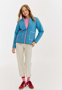 Niebieska pikowana kurtka z różowymi akcentami, dwiema kieszeniami naszytymi i zamkiem z przodu. Nosi się ją na różowej koszulce, w połączeniu z kremowymi spodniami i czerwonymi trampkami.