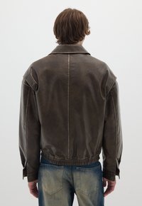 Giacca di pelle marrone con vestibilità comoda, ampio colletto, cinturino elastico in vita, cuciture dettagliate e maniche lunghe abbinata a jeans blu.
