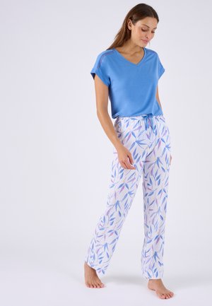 Mujer de pie descalza con una camiseta azul de manga corta y pantalones blancos con un patrón de hojas azul y morado sobre un fondo claro.