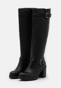 Bottes noires montantes jusqu'au genou en matériau synthétique lisse. Dotées d'une fermeture éclair latérale, de brides à boucles et d'un talon haut épais.